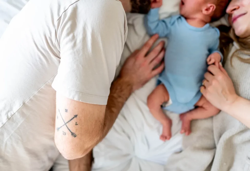 Eltern liegen mit ihrem Baby in hellen, neutralen Outfits auf dem Bett