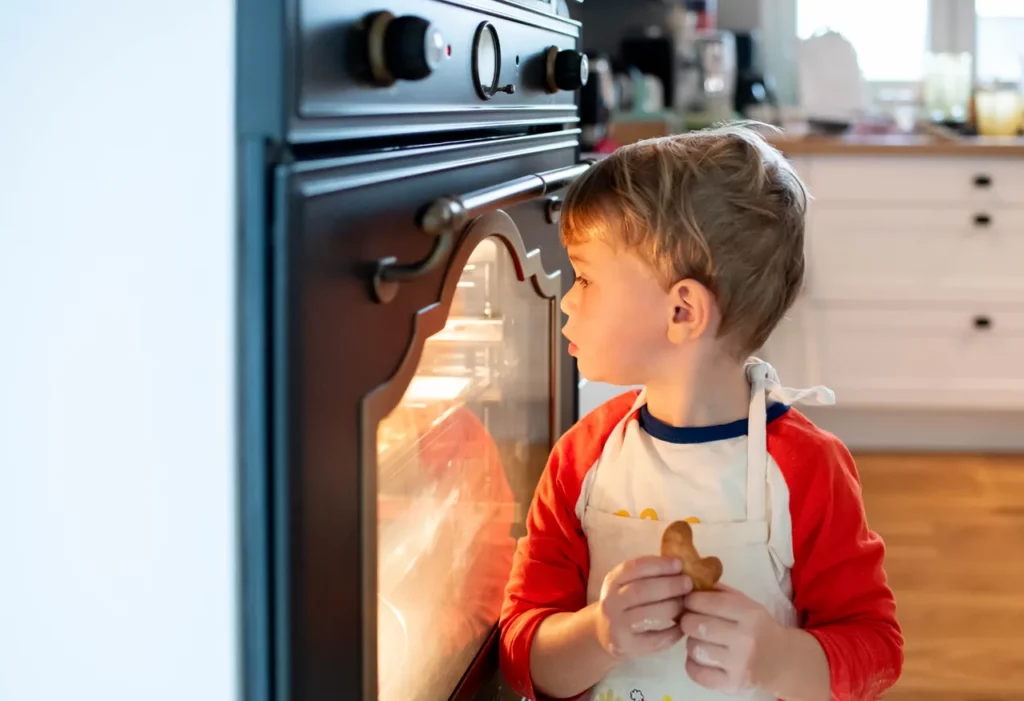 Kind mit Schürze und Plätzchen vor dem beleuchteten Backofen